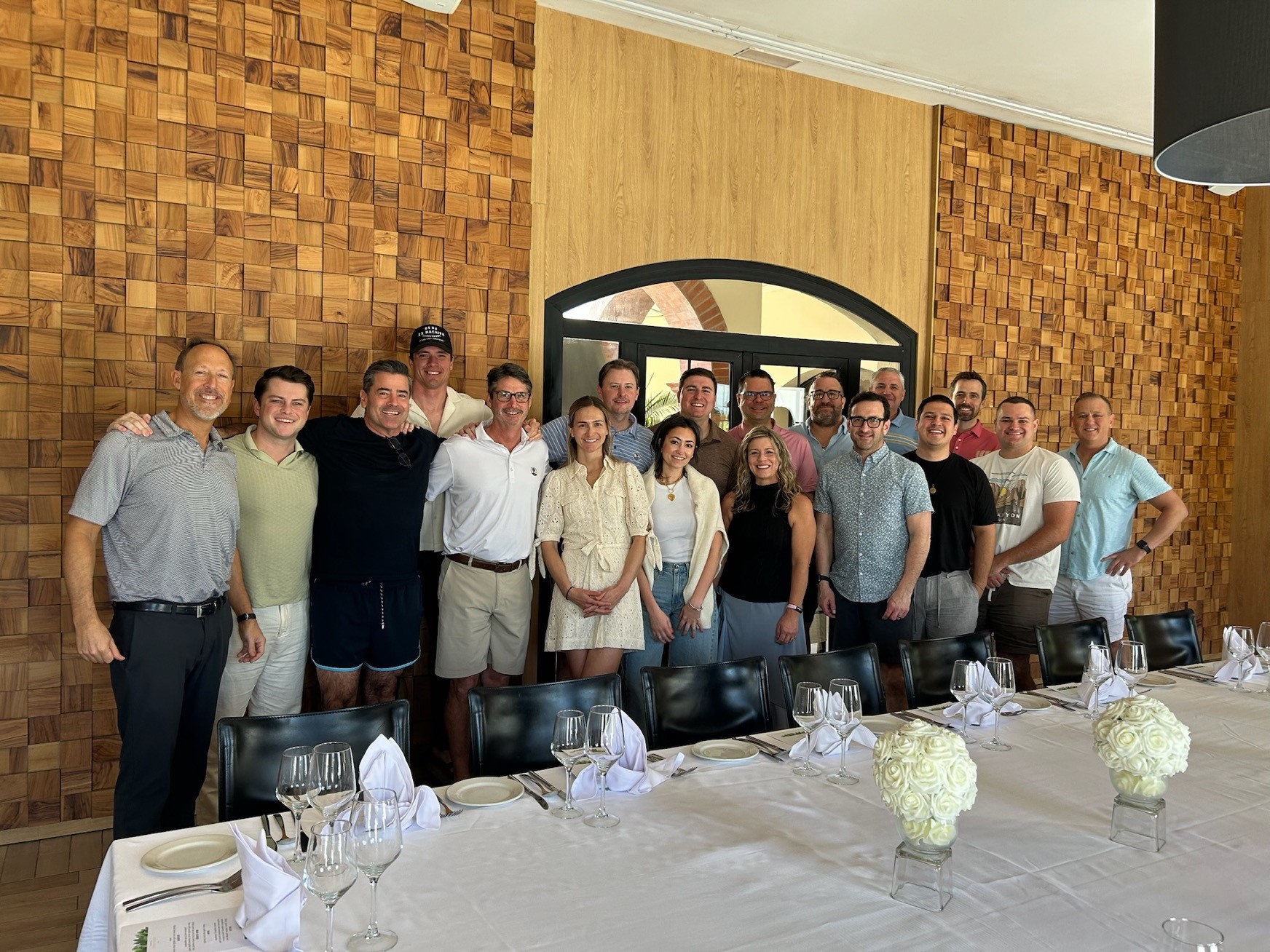 Team of sixteen employees from Gravity IT Resources smiling in a dining room with a wooden textured wall, set for a formal meal during a company incentive trip to the Dominican Republic. The cheerful and friendly atmosphere highlights how Gravity celebrates employee success and fosters a positive, rewarding company culture.
