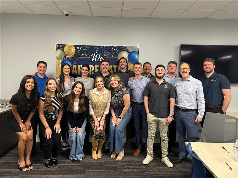 Team of sixteen employees at Gravity IT Resources smiling in a cheerful office setting with a festive banner and balloons in the background, showing how Gravity fosters a positive and appreciative company culture, making it a great place to work.